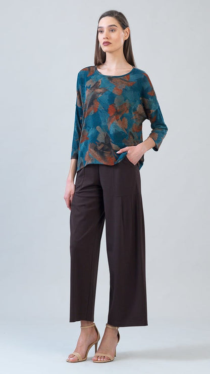 A woman modeling a long-sleeve blouse with a teal, brown, and rust abstract floral pattern, paired with dark brown wide-leg cropped pants and beige high-heeled sandals, against a light gray studio background.