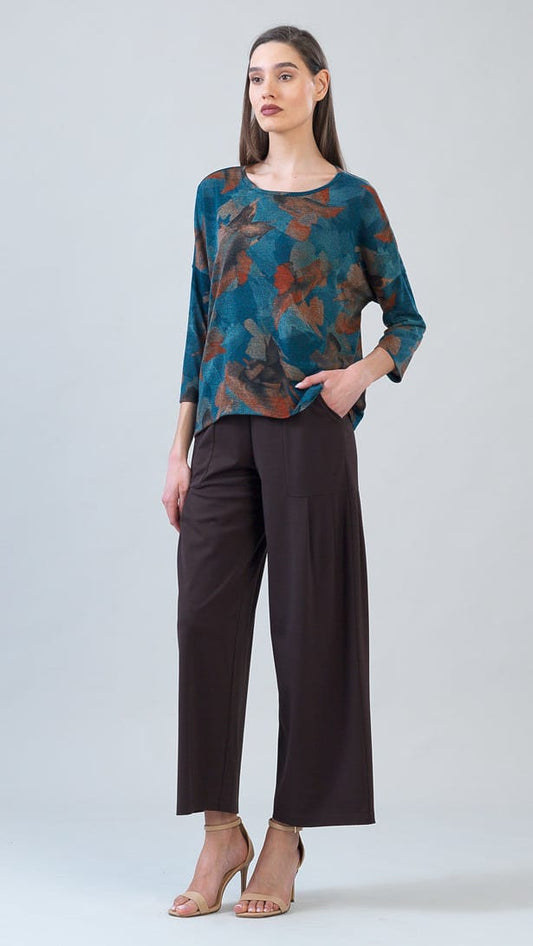 A woman modeling a long-sleeve blouse with a teal, brown, and rust abstract floral pattern, paired with dark brown wide-leg cropped pants and beige high-heeled sandals, against a light gray studio background.