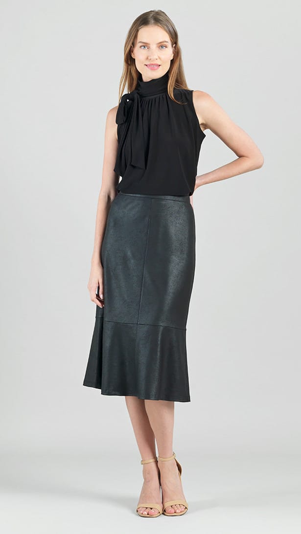 A woman modeling a sleeveless black mock-neck top with a large bow detail on the chest and a black faux-leather midi skirt with a ruffled hem, paired with beige high-heeled sandals, against a light gray studio background.