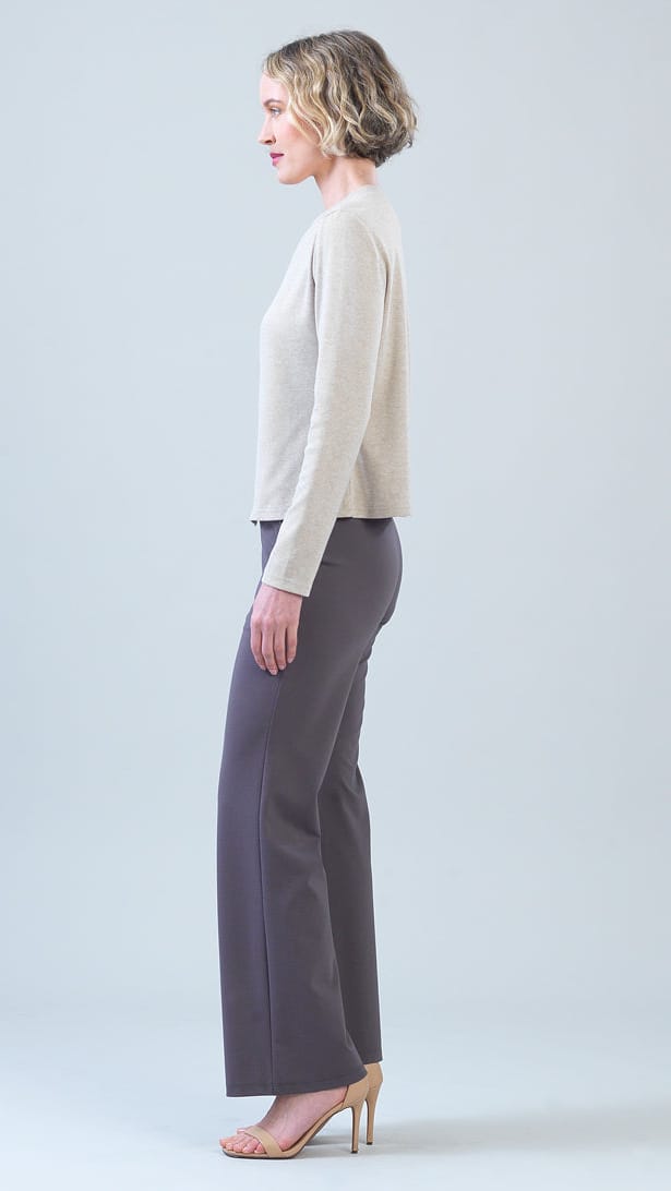 A woman in side profile wearing a light beige long-sleeve knit top, dark gray wide-leg trousers, and nude high heels, standing against a light gray studio background.