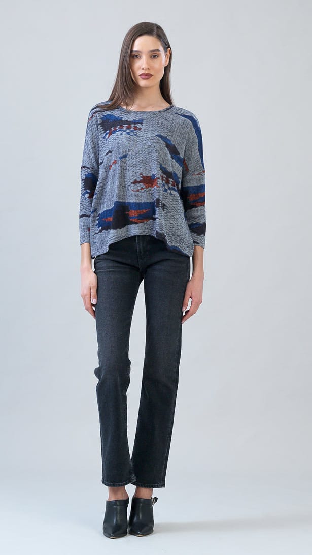 A woman stands against a light gray studio background wearing a loose, long-sleeve knit top with an abstract blue, red, and gray pattern, paired with dark charcoal jeans and black ankle boots.