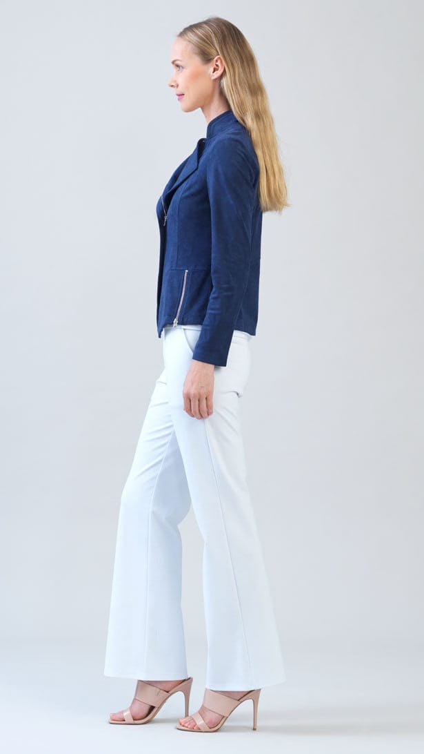 A woman stands in profile wearing a navy blue moto-style jacket, white wide-leg pants, and nude high-heeled sandals against a light gray studio background.