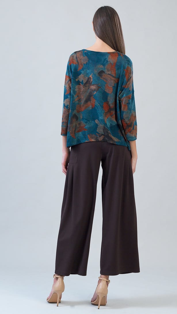 A woman is shown from the back wearing a three-quarter sleeve top with a teal and brown abstract floral pattern, paired with dark brown wide-leg pants and beige high-heeled sandals, set against a plain light gray studio background.
