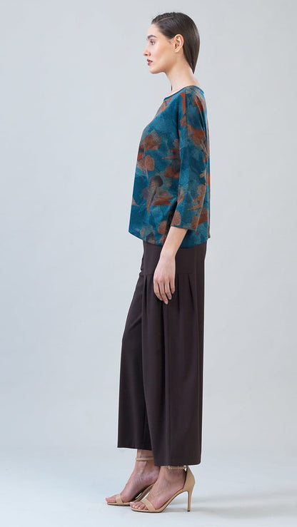 A woman stands in profile wearing a teal and rust abstract-print blouse with three-quarter sleeves and dark brown wide-leg cropped pants, paired with nude high-heeled sandals, against a light gray studio background.