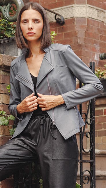 A woman models a gray zip-front jacket and black satin-like jogger pants with elastic cuffs, posing on outdoor brick steps against a red brick wall.