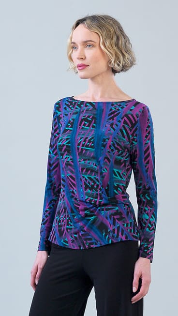 A woman modeling a long-sleeve boat-neck top with a vibrant abstract geometric pattern in purple, blue, and teal, paired with black pants against a light gray studio background.