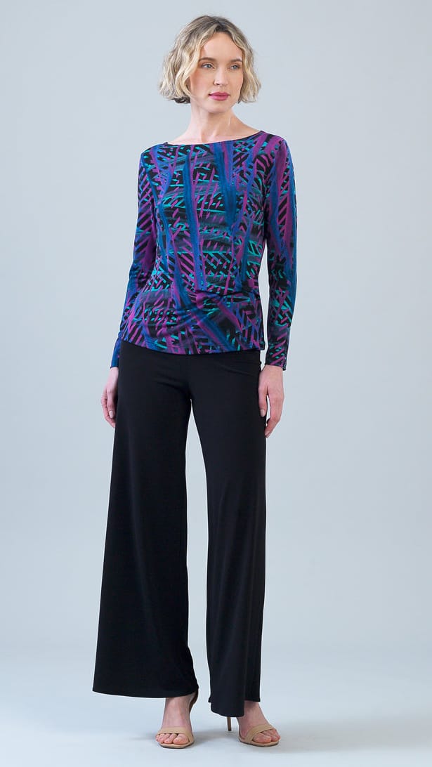 A woman modeling a long-sleeve boat-neck top with a vibrant purple, blue, and green abstract pattern, paired with black wide-leg trousers, against a light gray studio background.