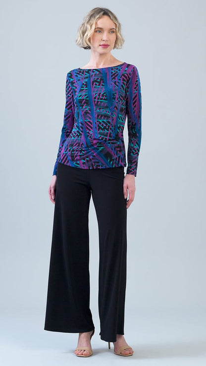 A woman modeling a long-sleeve boat-neck top with a vibrant purple, blue, and green abstract pattern, paired with black wide-leg trousers, against a light gray studio background.