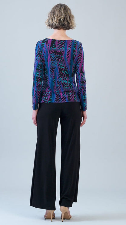 A woman viewed from the back wearing a long-sleeve boat-neck top with a vibrant blue, purple, and black abstract pattern, paired with black wide-leg trousers and beige heels, set against a light gray studio background.