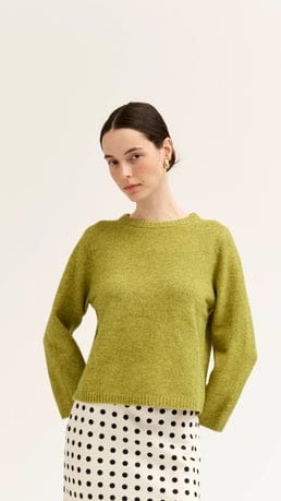 A woman wearing a cropped, long-sleeve knit sweater in a muted olive green, paired with a white skirt featuring black polka dots, set against a plain off-white studio background.
