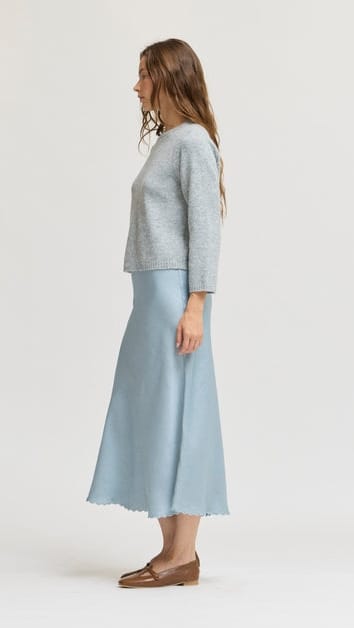 A woman stands in profile wearing a light gray three-quarter sleeve knit top and a light blue midi skirt with a raw hem, paired with brown leather loafers, against a plain light gray studio background.