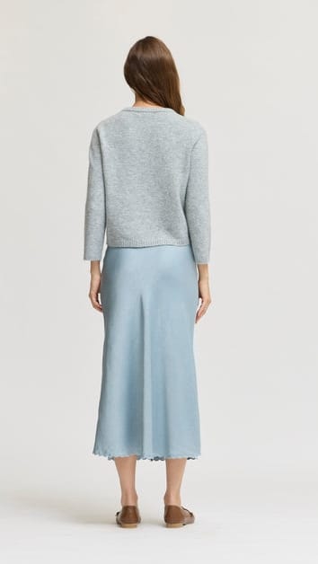 A woman viewed from the back wearing a light gray long-sleeve knit top and a light blue midi skirt, standing against a plain light gray studio background.