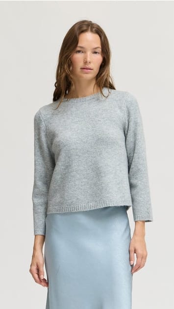 A woman wearing a light gray knit crewneck sweater and a light blue satin skirt, standing against a plain light gray studio background.