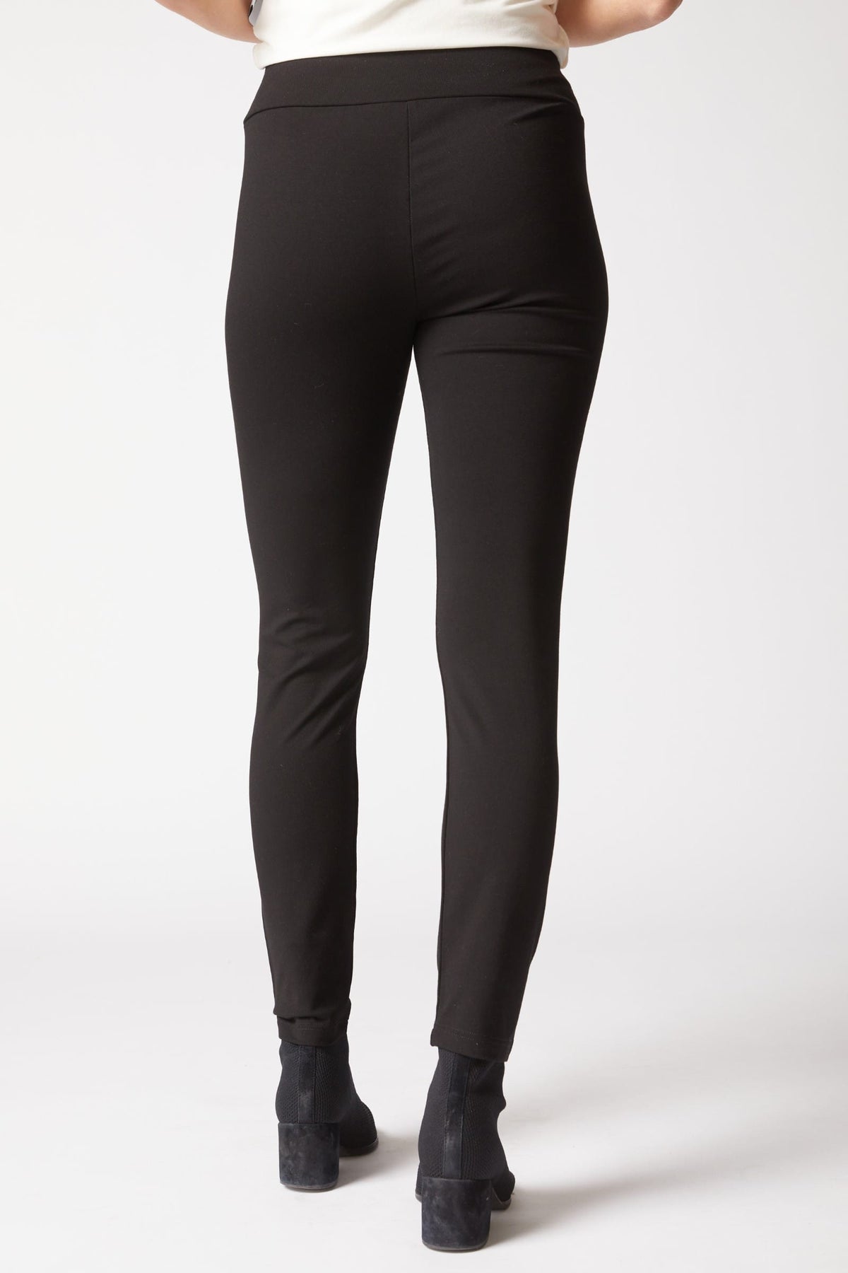 Rear view of a person wearing black high-waisted leggings and black ankle boots against a light gray studio background.