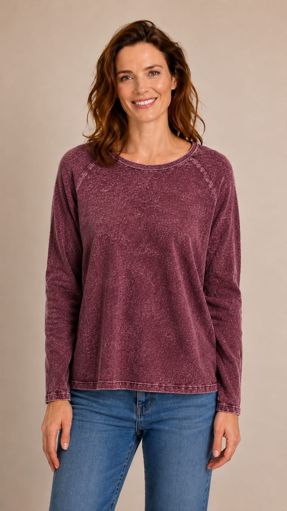 A woman wearing a long-sleeve, crew-neck, washed mauve top and blue jeans, standing against a solid beige studio background.