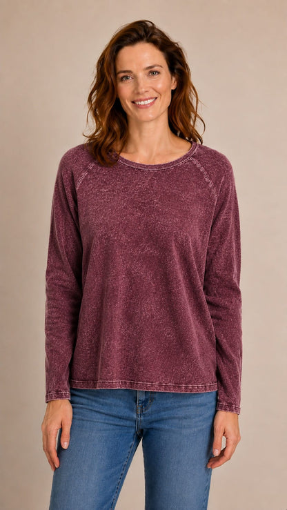 A woman wearing a long-sleeve, crew-neck, washed mauve top and blue jeans, standing against a solid beige studio background.