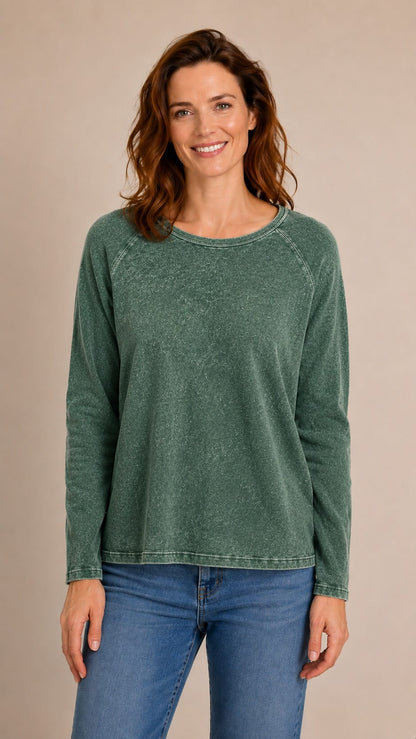 A woman wearing a long-sleeve crewneck top in a washed forest green color, paired with blue jeans, standing against a solid beige studio background.