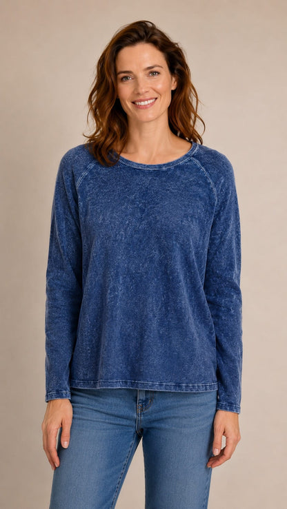 A woman wearing a long-sleeve, crew-neck top in a washed blue denim-like fabric, paired with blue jeans, standing against a solid beige studio background.