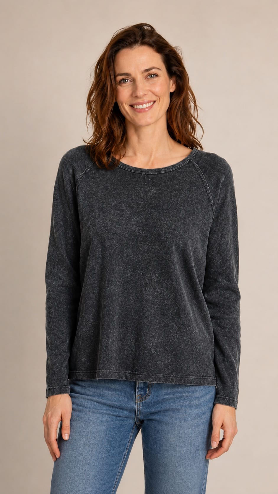 A woman wearing a dark charcoal gray long-sleeve crewneck sweater and blue jeans, standing against a plain beige studio background.