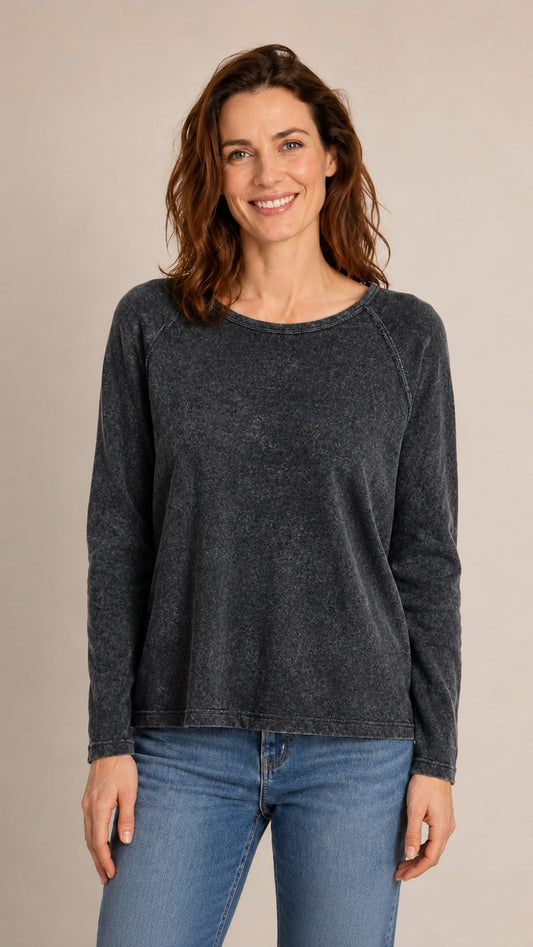 A woman wearing a dark charcoal gray long-sleeve crewneck sweater and blue jeans, standing against a plain beige studio background.