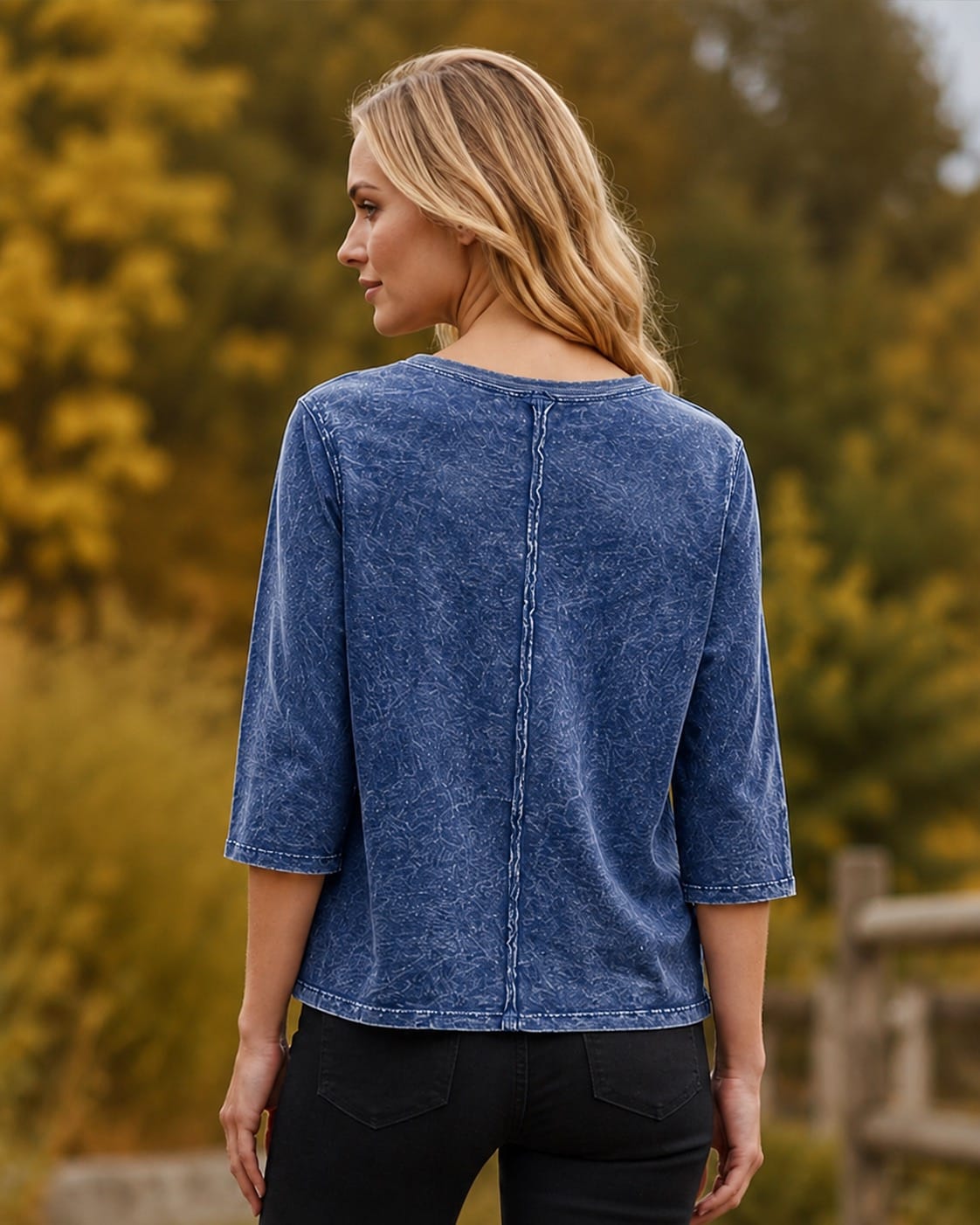 A woman shown from the back wearing a blue washed three-quarter sleeve top and black pants, standing outdoors with a blurred background of green and yellow foliage.