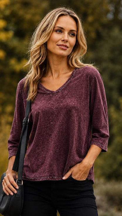 A woman wearing a maroon three-quarter sleeve V-neck top with a washed texture, paired with black pants and carrying a black shoulder bag, set against a blurred outdoor background of green and yellow foliage.