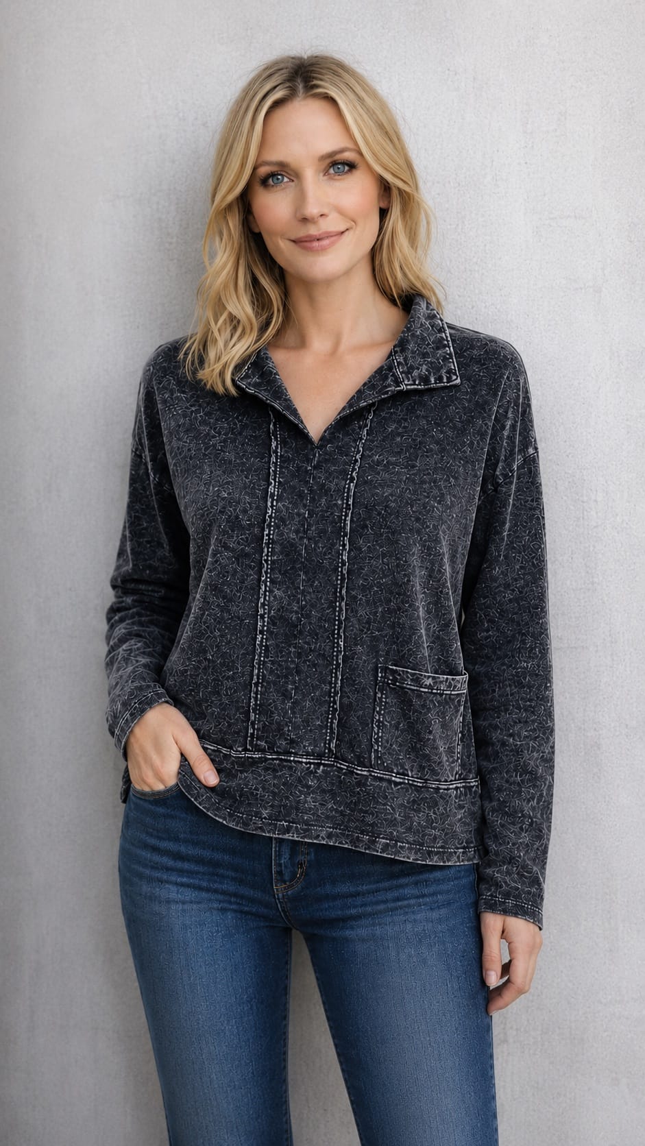 A woman wearing a dark charcoal gray, washed-texture long-sleeve quarter-zip pullover with a stand collar and front patch pockets, paired with blue jeans, standing against a textured gray wall.