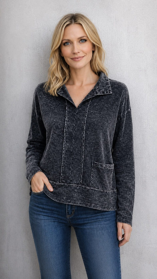A woman wearing a dark charcoal gray, washed-texture long-sleeve quarter-zip pullover with a stand collar and front patch pockets, paired with blue jeans, standing against a textured gray wall.
