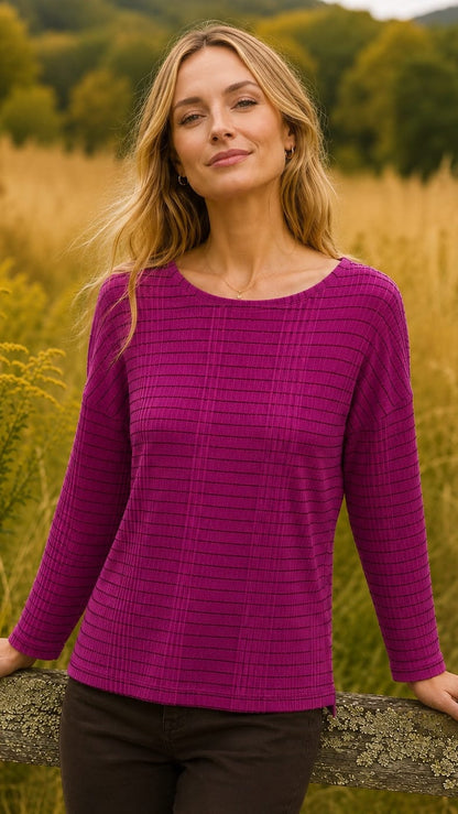 A woman wearing a textured magenta long-sleeve top and dark brown pants, standing outdoors in a field of golden grass.