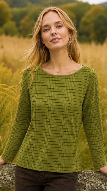 A woman wearing a textured olive green long-sleeve top with a subtle grid pattern, standing outdoors in a field of golden grass.