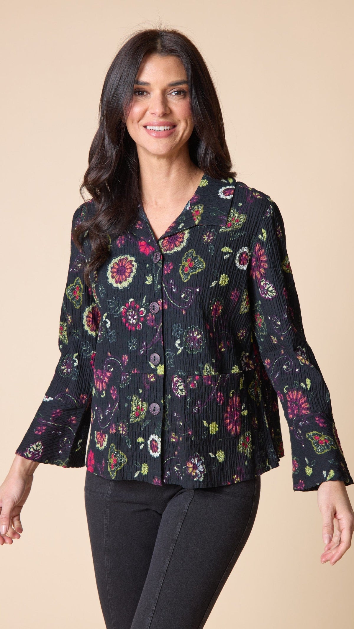 A woman modeling a black long-sleeve blouse with a colorful floral and paisley print, paired with black pants, against a solid beige background.
