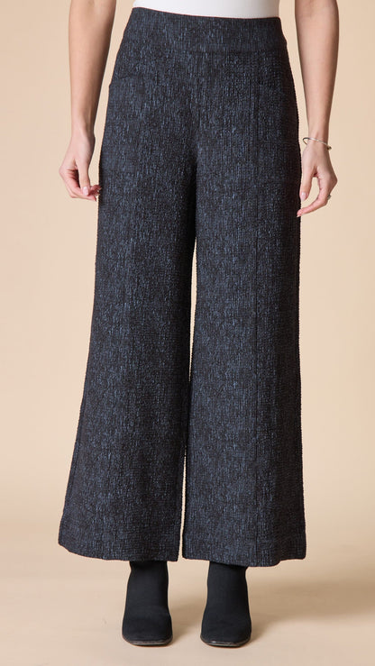 A person wearing dark charcoal wide-leg cropped pants with a textured tweed-like weave, paired with black ankle boots, against a solid beige studio background.