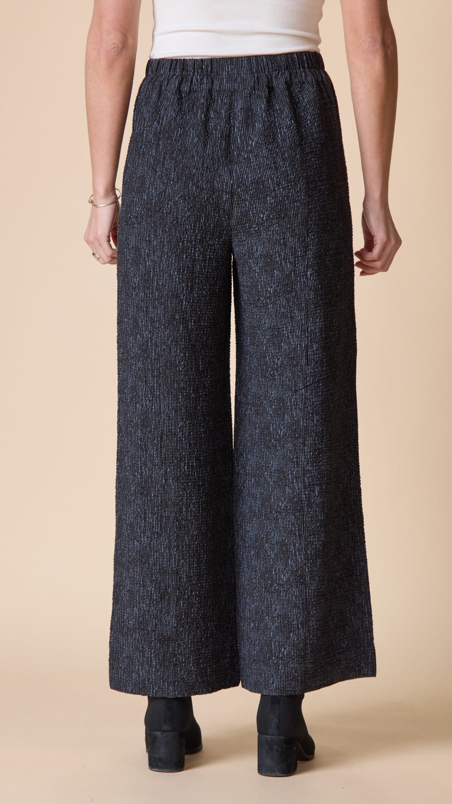 A person shown from the back wearing dark charcoal wide-leg cropped pants with a textured heathered pattern, paired with a white top and black ankle boots, against a solid beige studio background.