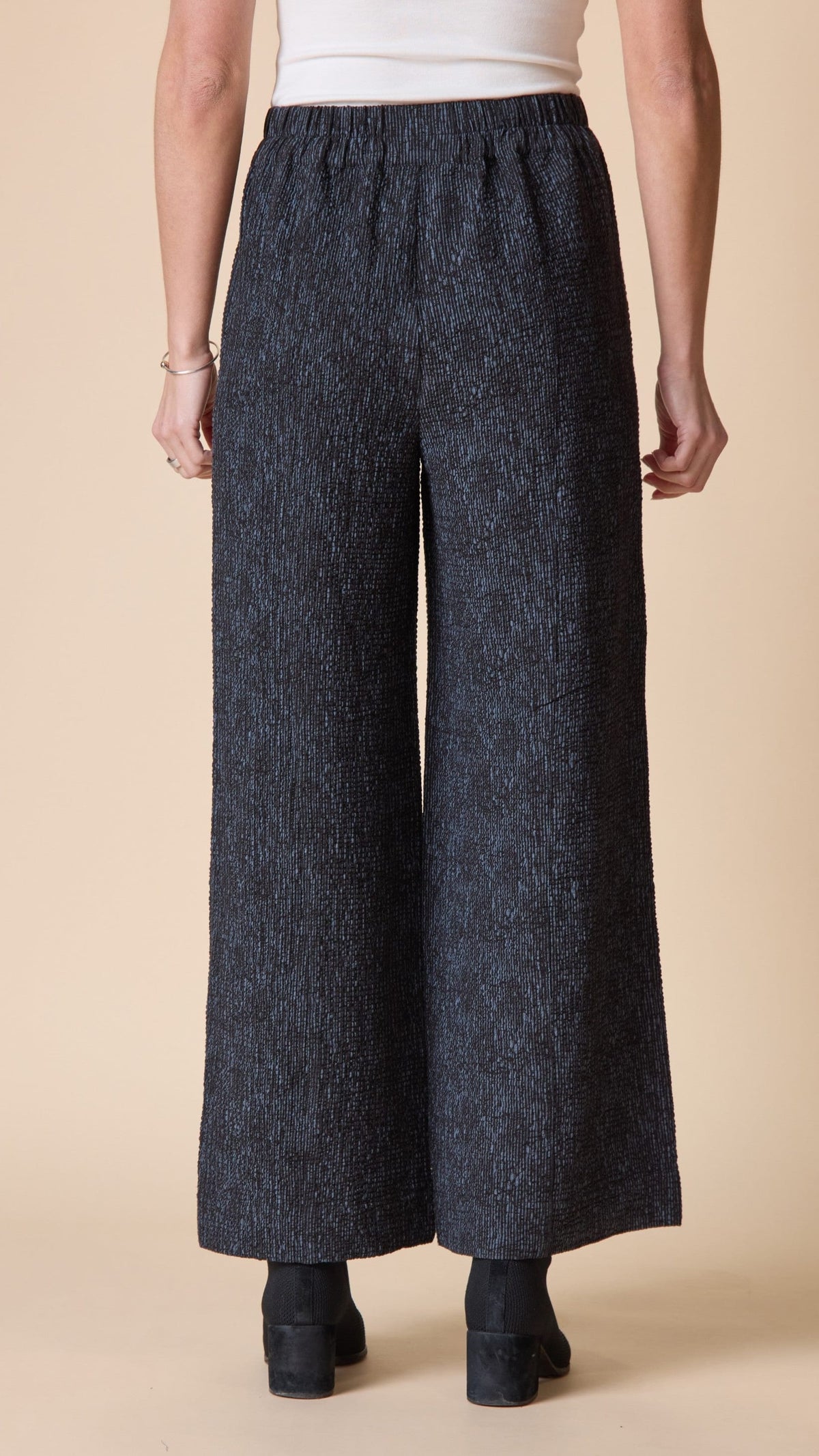 A person shown from the back wearing dark charcoal wide-leg cropped pants with a textured heathered pattern, paired with a white top and black ankle boots, against a solid beige studio background.