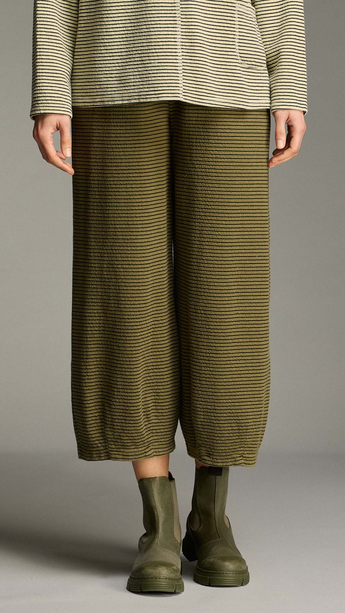 A model is shown from the waist down wearing olive green and black horizontally striped wide-leg cropped pants, a matching striped top, and olive green Chelsea boots against a neutral gray studio background.