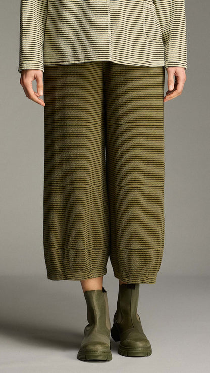 A model is shown from the waist down wearing olive green and black horizontally striped wide-leg cropped pants, a matching striped top, and olive green Chelsea boots against a neutral gray studio background.