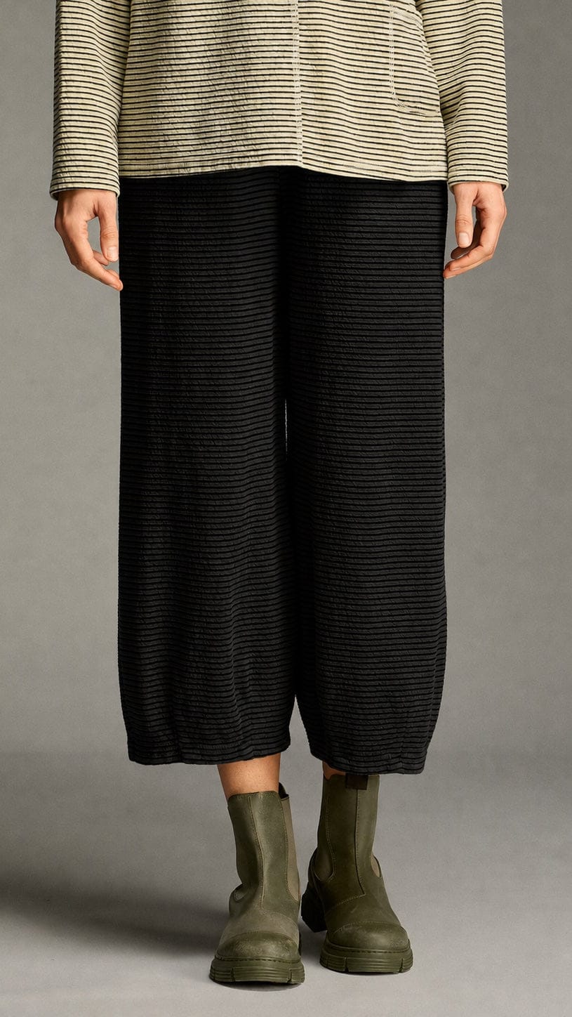 A model is shown from the waist down wearing black wide-leg cropped trousers with fine horizontal stripes, paired with olive green Chelsea boots and a beige and black striped long-sleeve top, set against a neutral gray studio background.