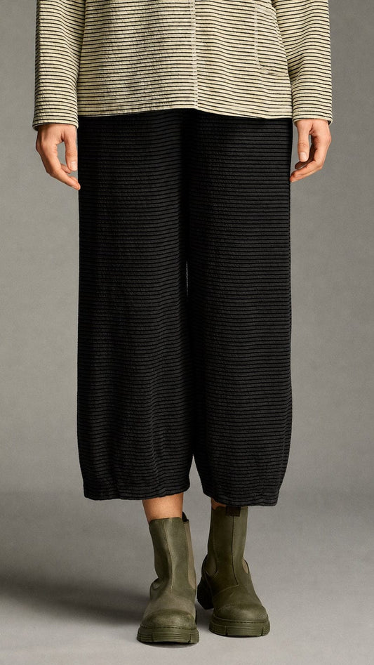 A model is shown from the waist down wearing black wide-leg cropped trousers with fine horizontal stripes, paired with olive green Chelsea boots and a beige and black striped long-sleeve top, set against a neutral gray studio background.