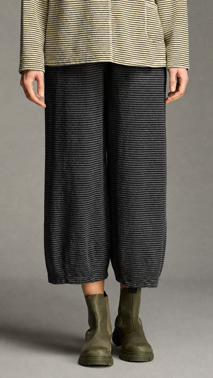 A person wearing loose, wide-leg cropped pants in a dark charcoal gray and black striped knit, paired with olive green Chelsea boots and a beige-and-gray striped top, photographed against a neutral gray studio background.