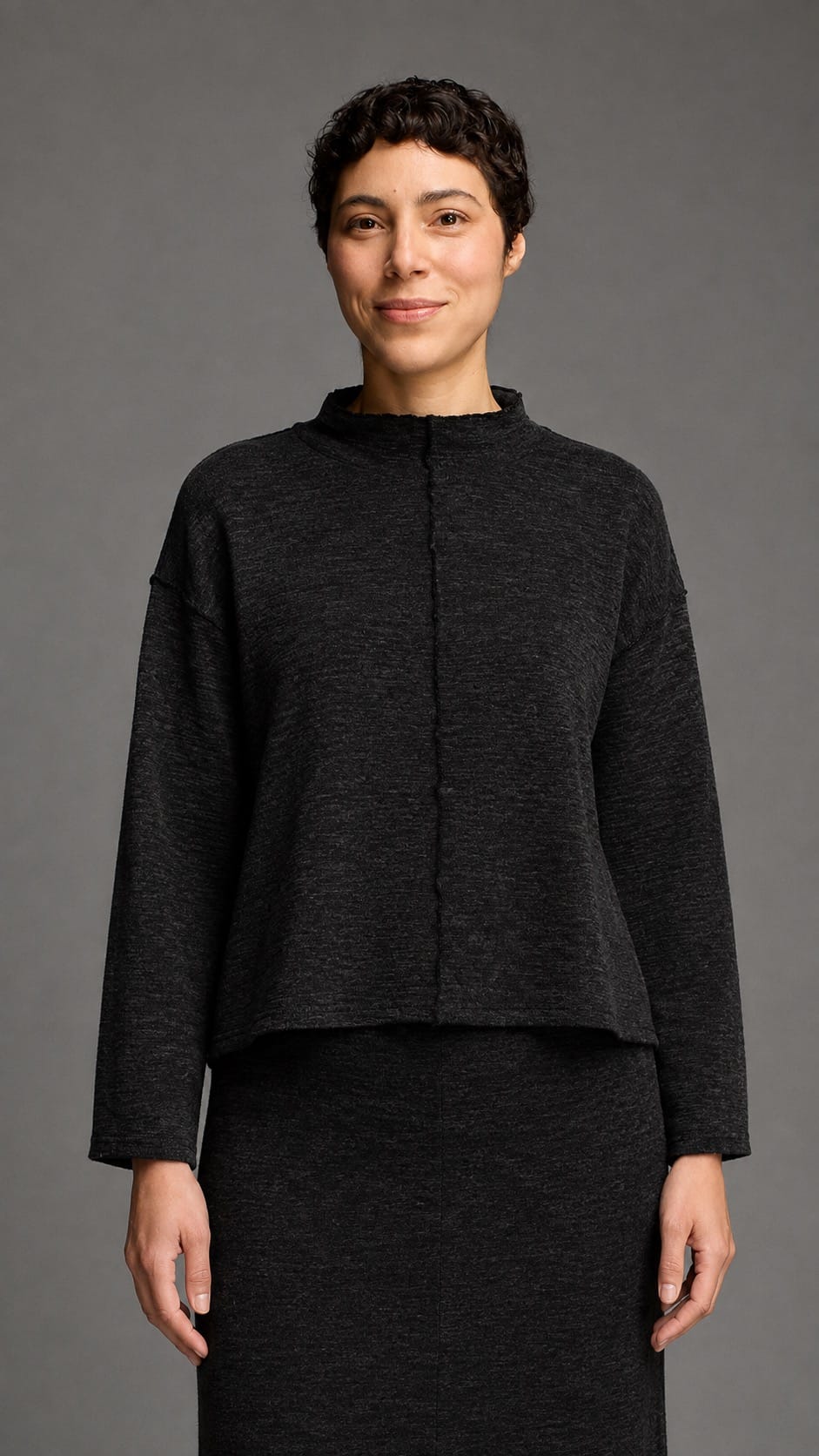 A model wearing a dark charcoal gray cropped knit top with a mock neck and front button closure, paired with a matching dark skirt, against a solid gray studio background.