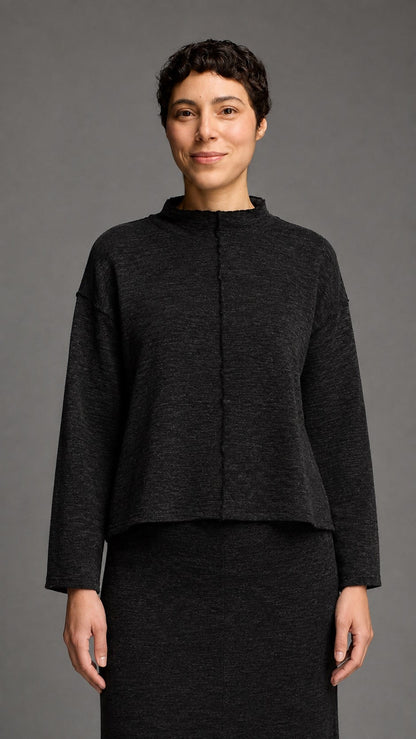 A model wearing a dark charcoal gray cropped knit top with a mock neck and front button closure, paired with a matching dark skirt, against a solid gray studio background.