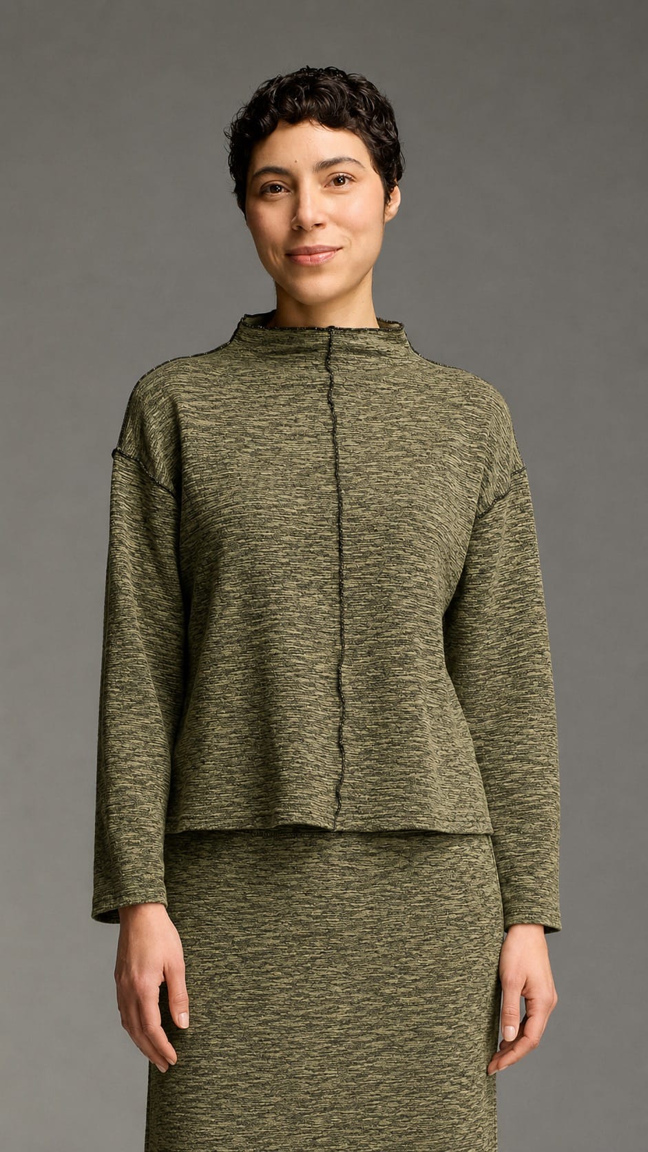 A model wearing a matching olive green knit two-piece set consisting of a cropped long-sleeve top and a high-waisted skirt, photographed against a solid gray studio background.