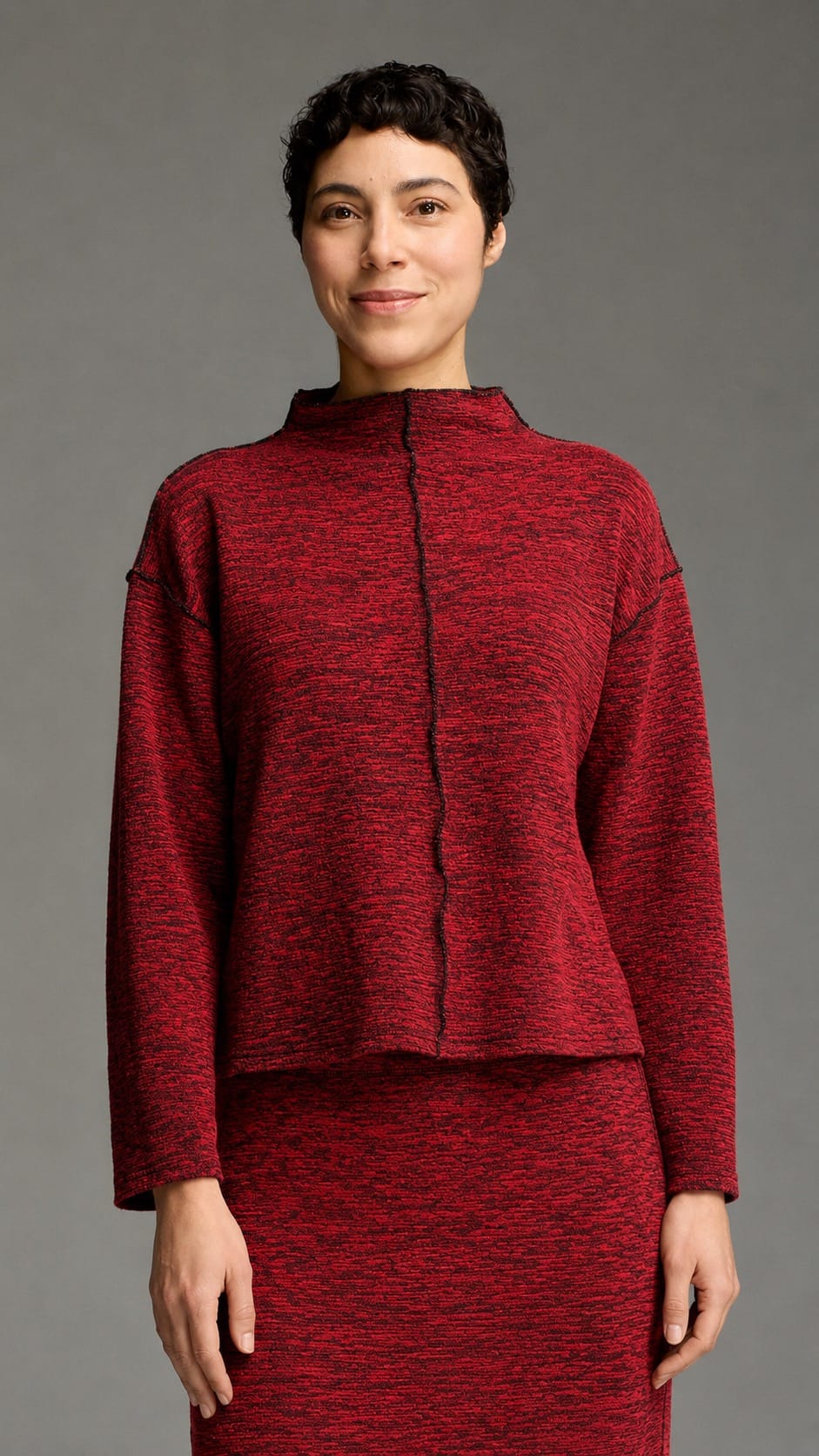 A model wearing a textured red and black knit two-piece set consisting of a cropped long-sleeve top with a mock neck and a matching skirt, shot against a solid gray studio background.