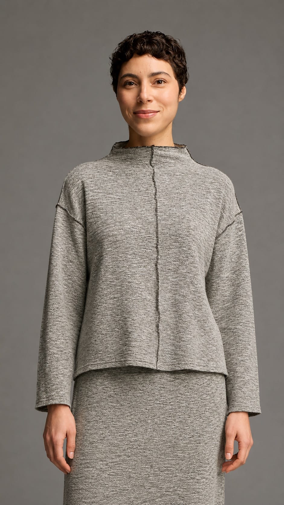 A woman modeling a gray heathered knit two-piece outfit consisting of a cropped long-sleeve top with a mock neck and a matching high-waisted skirt, against a solid gray studio background.