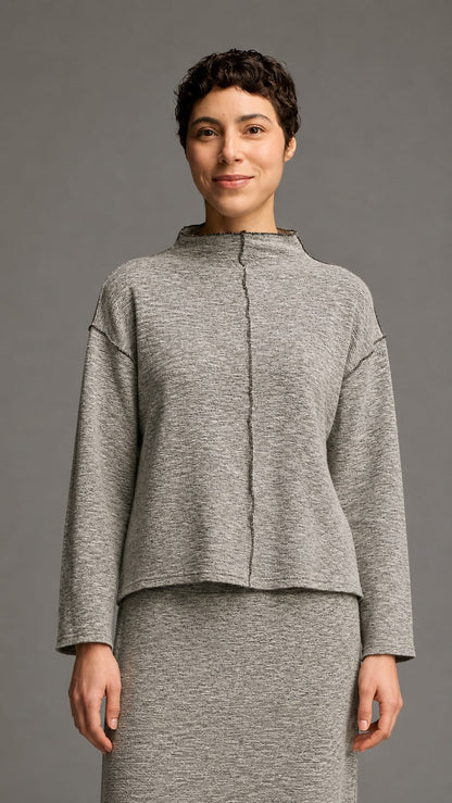 A woman modeling a gray heathered knit two-piece outfit consisting of a cropped long-sleeve top with a mock neck and a matching high-waisted skirt, against a solid gray studio background.