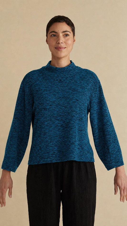 A woman modeling a long-sleeve mock neck knit sweater in a heathered teal blue color, paired with black pants, against a solid beige background.