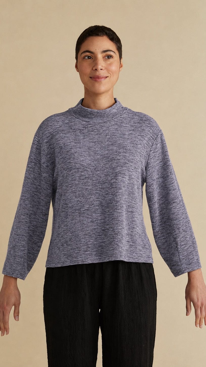 A woman modeling a heathered slate blue mock neck long-sleeve top with black pleated trousers, standing against a solid beige background.
