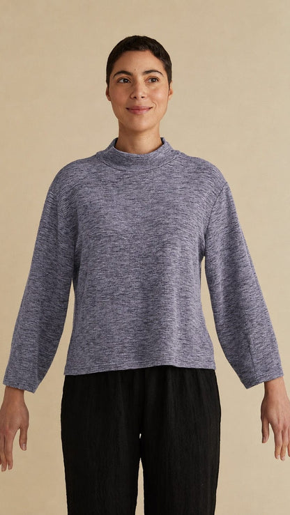 A woman modeling a heathered slate blue mock neck long-sleeve top with black pleated trousers, standing against a solid beige background.