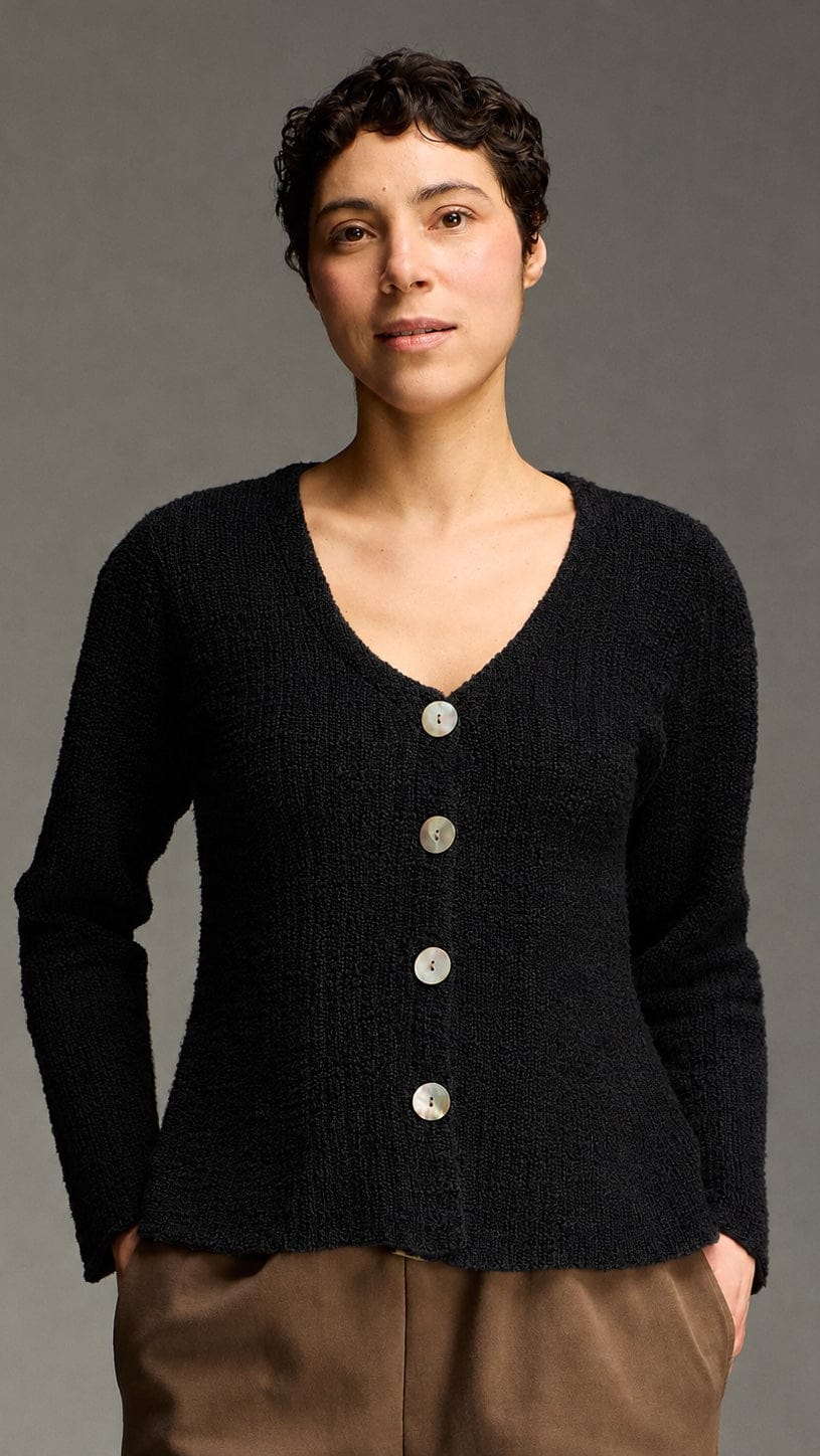 A model wearing a black textured knit cardigan with a V-neck and four silver buttons, paired with brown pants, against a solid gray studio background.