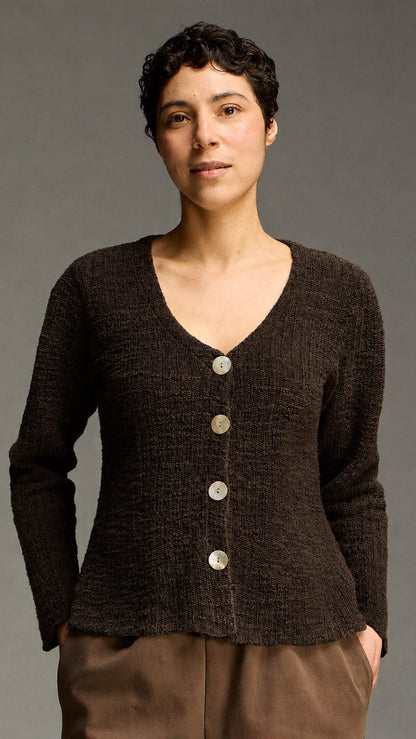 A woman wearing a dark brown textured knit cardigan with a V-neck and four large buttons, paired with brown pants, photographed against a neutral gray studio background.